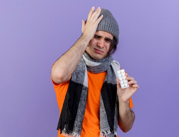 Bezpłatne zdjęcie niezadowolony młody chory w czapce zimowej z szalikiem trzymając pigułki kładąc rękę na bolącej głowie na białym tle na fioletowym tle