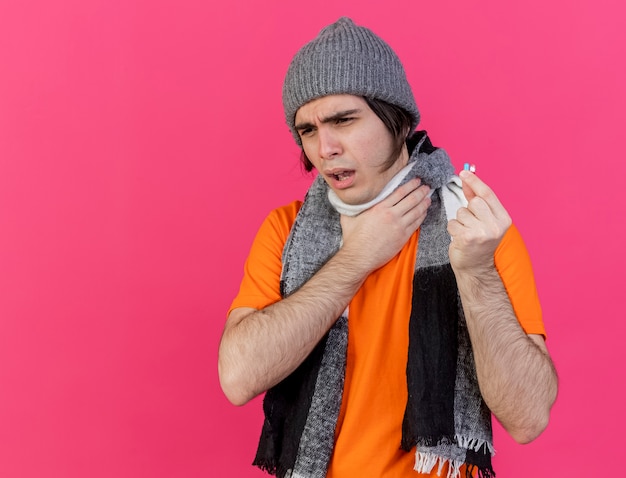 Bezpłatne zdjęcie niezadowolony młody chory w czapce zimowej z szalikiem trzymającym pigułkę i złapał bolące gardło odizolowane na różowo
