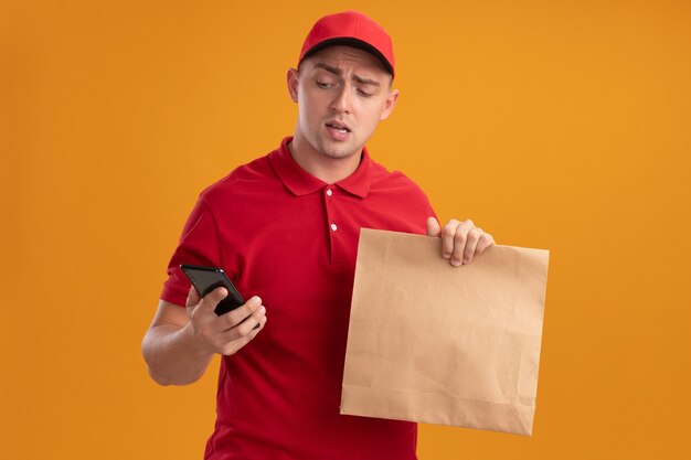 Niezadowolony młody człowiek dostawy ubrany w mundur z czapką, trzymając papierowy pakiet żywności, patrząc na telefon w ręku na białym tle na pomarańczowej ścianie