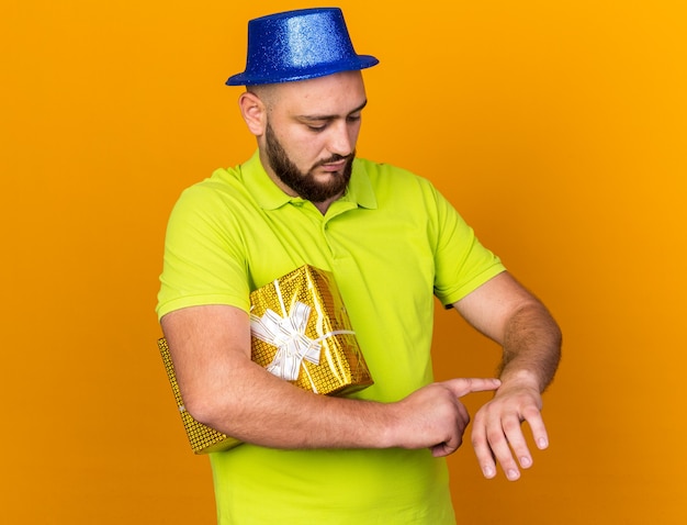 Niezadowolony młody człowiek w kapeluszu imprezowym, trzymający pudełko pokazujące gest zegara na nadgarstku na pomarańczowej ścianie