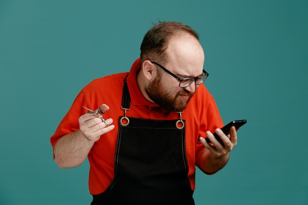 Niezadowolony młody fryzjer męski w okularach, czerwona koszula i fartuch fryzjerski, trzymając nożyczki i telefon komórkowy, patrząc na telefon na białym tle na niebieskim tle