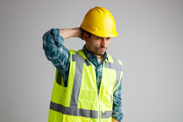 Niezadowolony młody inżynier w kasku ochronnym i mundurze, patrząc na bok, trzymając rękę z tyłu głowy z bólem głowy na białym tle