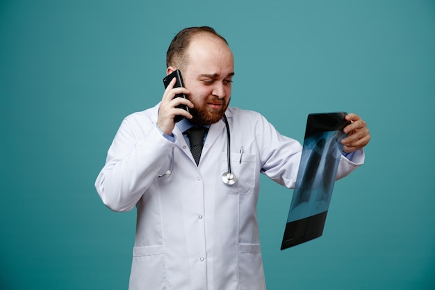 Niezadowolony młody lekarz mężczyzna ubrany w fartuch medyczny i stetoskop na szyi, trzymając i patrząc na zdjęcie rentgenowskie podczas rozmowy przez telefon na białym tle na niebieskim tle