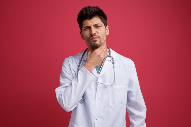 Niezadowolony młody lekarz mężczyzna ubrany w mundur medyczny i stetoskop na szyi, trzymając rękę na szyi, patrząc na kamerę mającą ból gardła na białym tle na czerwonym tle