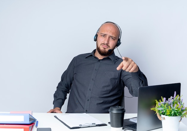 Niezadowolony młody łysy mężczyzna call center sobie zestaw słuchawkowy siedzi przy biurku z narzędzi pracy, wskazując na aparat na białym tle