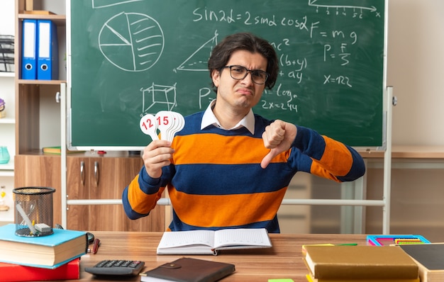 Bezpłatne zdjęcie niezadowolony młody nauczyciel geometrii w okularach siedzący przy biurku z przyborami szkolnymi w klasie, patrzący na przód pokazujący wachlarze liczbowe i kciuk w dół