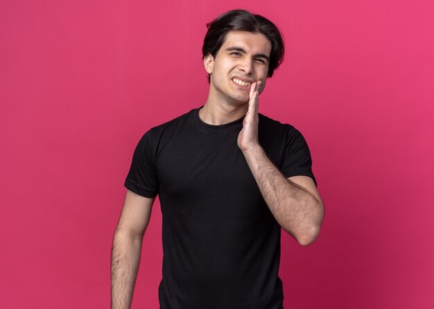 Niezadowolony młody przystojny facet ubrany w czarną koszulkę, kładący rękę na bolącym zębie odizolowanym na różowej ścianie