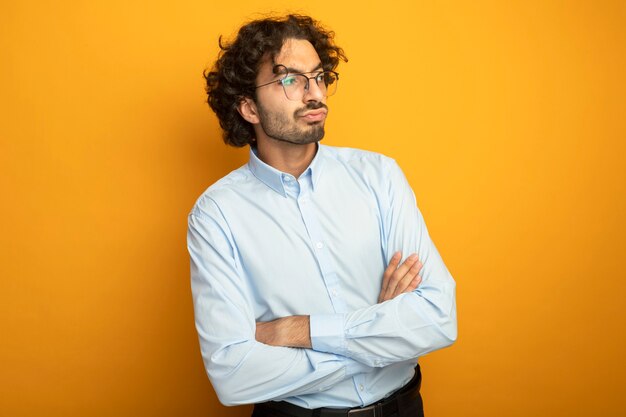 Niezadowolony młody przystojny kaukaski mężczyzna w okularach stojący z zamkniętą postawą, patrząc z boku na białym tle na pomarańczowym tle z miejsca na kopię