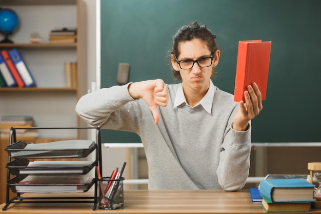 niezadowolony pokazując kciuk w dół młody nauczyciel płci męskiej trzymający książkę siedzącą przy biurku z włączonymi narzędziami szkolnymi w klasie