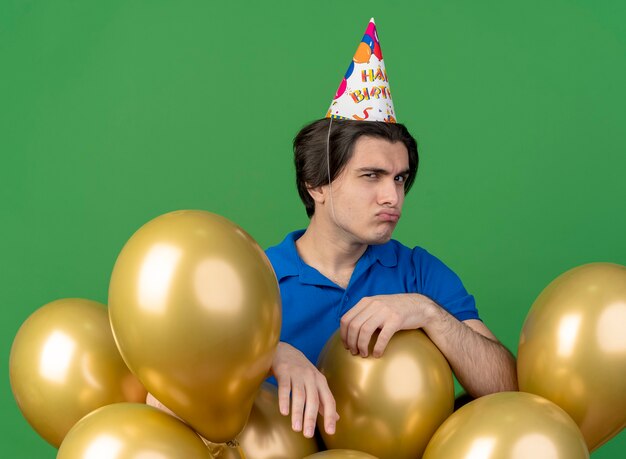 Niezadowolony przystojny kaukaski mężczyzna w czapce urodzinowej stoi z balonami z helem, patrząc na kamerę