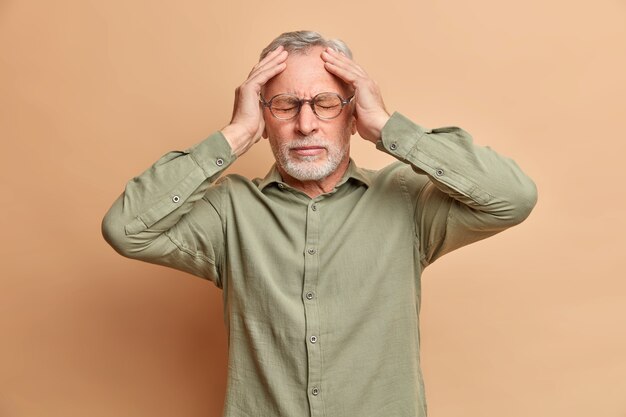 Niezadowolony siwy mężczyzna cierpi na ból głowy trzyma ręce na głowie, aby ujawnić potrzebę bólu środki przeciwbólowe ma migrenę po hałaśliwej imprezie nosi formalną koszulę odizolowaną na brązowej ścianie