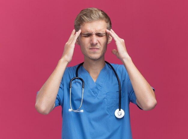Niezadowolony z zamkniętymi oczami młody lekarz w mundurze lekarza ze stetoskopem kładzie palce na bolących skroniach na różowej ścianie