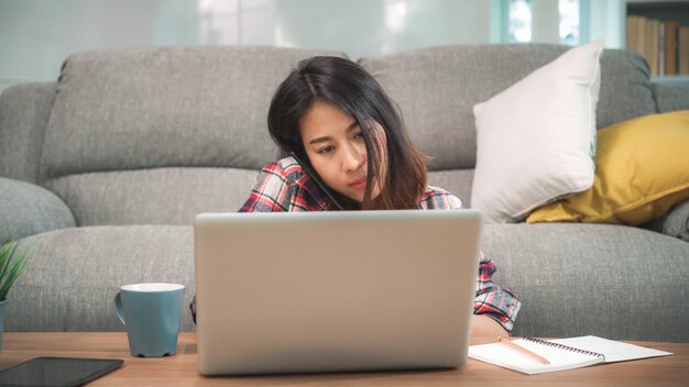 Niezależna Azjatycka kobieta pracuje w domu, biznesowa kobieta pracuje na laptopie i używa telefon komórkowego opowiada z klientem na kanapie w żywym pokoju w domu.