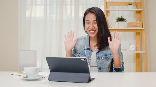 Niezależna biznesowych kobiet przypadkowa odzież używać pastylki działania wezwania wideokonferencję z klientem w miejscu pracy w żywym pokoju w domu. Szczęśliwa młoda Azjatycka dziewczyna relaksuje obsiadanie na biurku robi pracie w internecie.