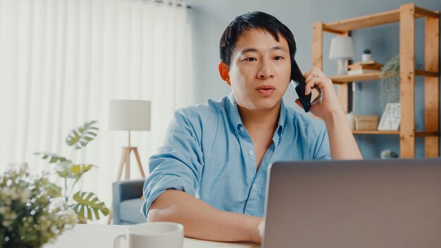 Niezależny facet z Azji na co dzień przy użyciu rozmowy laptopa na telefon komórkowy w salonie w domu.