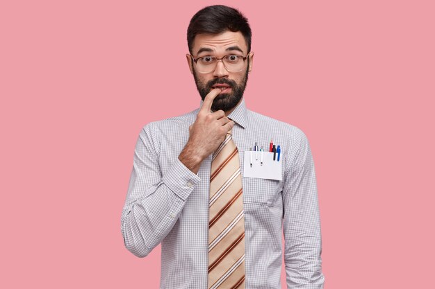 Niezdecydowany intelektualista trzyma palec przy ustach, ma gęstą brodę, ubrany w formalną koszulę i krawat, nosi duże okulary