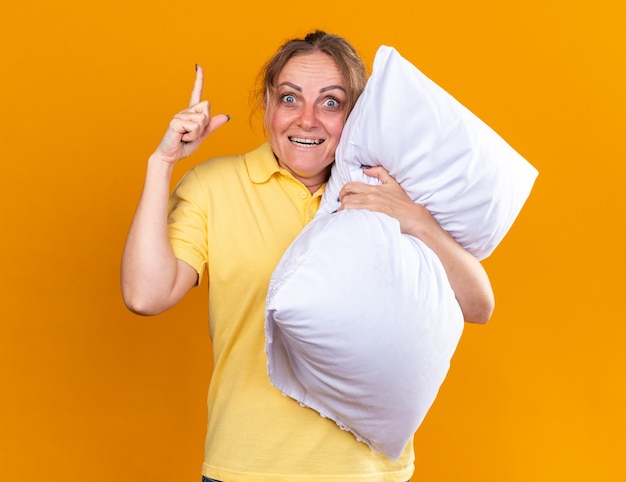 Niezdrowa kobieta w żółtej koszuli, cierpiąca na grypę i przeziębienie, przytula poduszkę pokazującą indeks fignera pokazujący indeks fignera uśmiechający się, lepiej stojący nad pomarańczową ścianą