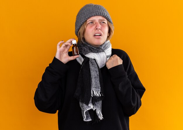 Niezdrowa młoda kobieta w ciepłej czapce z szalikiem na szyi mdłości cierpiąca na przeziębienie i grypę trzymająca butelkę z lekarstwem patrząc na bok ze zirytowanym wyrazem twarzy na pomarańczowej ścianie