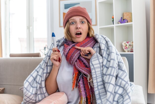 Niezdrowa młoda kobieta w ciepłym kapeluszu owinięta w koc, wyglądająca na chorą i chorą, cierpiącą na przeziębienie i grypę, trzymająca termometr, mająca gorączkę, wyglądającą na zmartwioną, siedzącą na kanapie w jasnym salonie