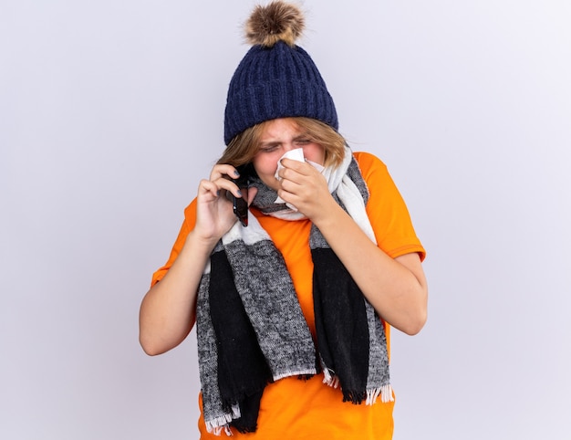 Bezpłatne zdjęcie niezdrowa młoda kobieta w pomarańczowym t-shircie z ciepłym szalikiem na szyi i kapeluszu czuje się okropnie rozmawiając przez telefon komórkowy dmuchając katar kichając w tkance stojącej nad białą ścianą