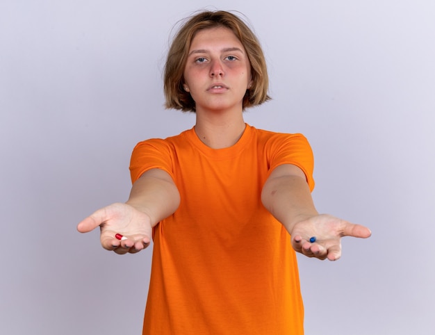 Bezpłatne zdjęcie niezdrowa młoda kobieta w pomarańczowym t-shircie źle się czuje, cierpi na grypę pokazując tabletki w dłoniach ze smutnym wyrazem twarzy stojącej nad białą ścianą