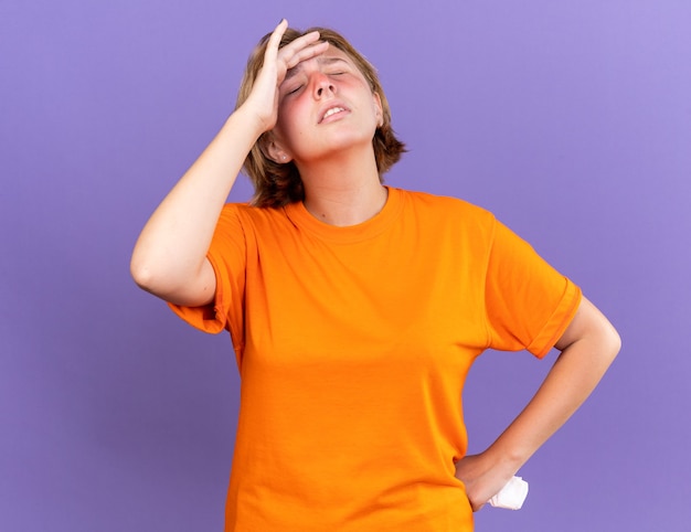 Niezdrowa młoda kobieta w pomarańczowym t-shircie źle się czuje dotykając czoła, mając zawroty głowy z grypą stojącą nad fioletową ścianą