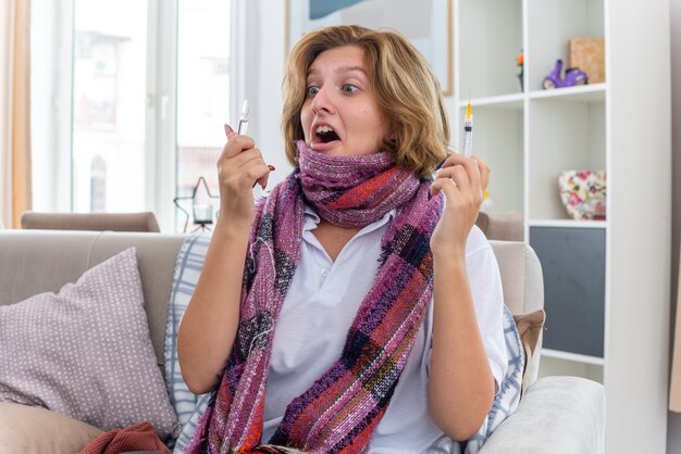 Bezpłatne zdjęcie niezdrowa młoda kobieta z ciepłym szalikiem na szyi źle się czuje i cierpi na przeziębienie i grypę trzymając strzykawkę i ampułkę wyglądającą na zmartwioną i zdziwioną siedzącą na kanapie w jasnym salonie
