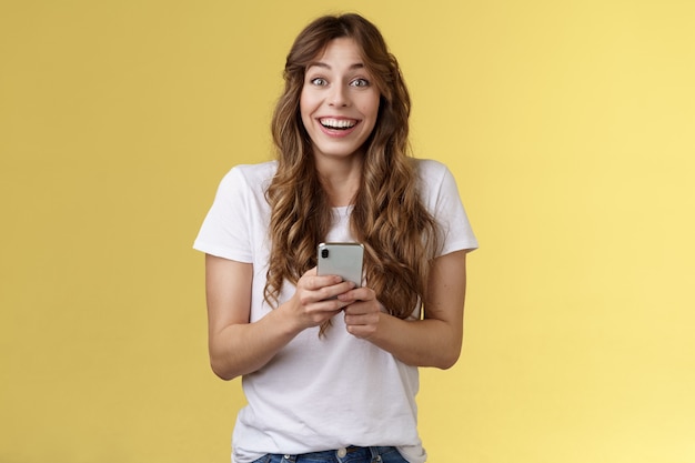 Niezwykle szczęśliwy entuzjastyczny zdziwiony radosny urocza kobieca dziewczyna otrzymuje przyjemną wzruszającą wiadomość online trzymaj smartfon uśmiechając się szeroko patrz aparat radosny rozbawiony żółtym tle