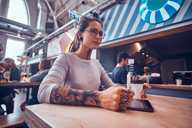 Niski kąt zdjęcie atrakcyjnej zamyślonej dziewczyny z dredami i tatuażami. Dhe szkicuje siedząc w kawiarni.