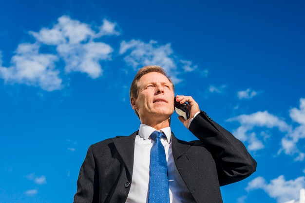 Bezpłatne zdjęcie niskiego kąta widok dojrzały biznesmen opowiada na telefonie komórkowym przeciw niebu