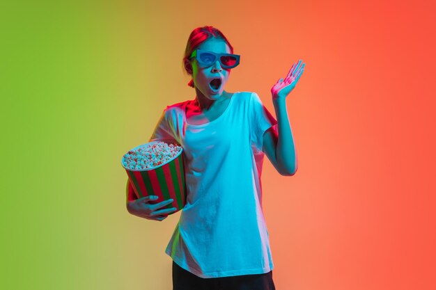 Noszenie zestawu słuchawkowego 3D zszokowany przestraszony popcornem Portret młodej dziewczyny rasy kaukaskiej na gradientowym zielono-pomarańczowym studio w tle w świetle neonowym Koncepcja młodych ludzkich emocji wyraz twarzy