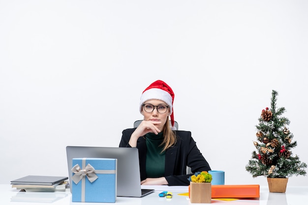 Noworoczny nastrój z młodą atrakcyjną kobietą w czapce Świętego Mikołaja siedzącą przy stole z choinką i prezentem i czekającą na biuro