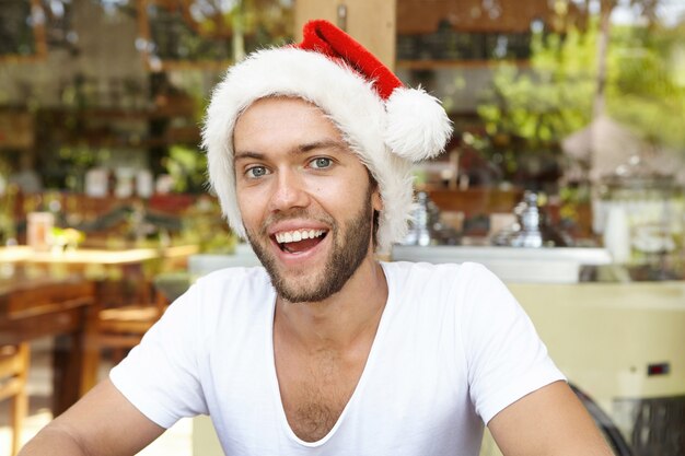Obchody Bożego Narodzenia i Nowego Roku. Kryty portret szczęśliwy atrakcyjny młody człowiek rasy kaukaskiej z brodą pozowanie w kawiarni na sobie czerwony kapelusz Świętego Mikołaja, śmiejąc się radośnie podczas wakacji