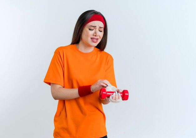 Obolała młoda sportowa kobieta nosząca opaskę i opaski na rękę trzymająca hantle dotykająca i patrząc na zraniony nadgarstek owinięty bandażem na białym tle