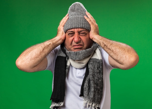 Bezpłatne zdjęcie obolały dorosły chory kaukaski mężczyzna z szalikiem na szyi w czapce zimowej trzymającej głowę odizolowaną na zielonej ścianie z kopią przestrzeni