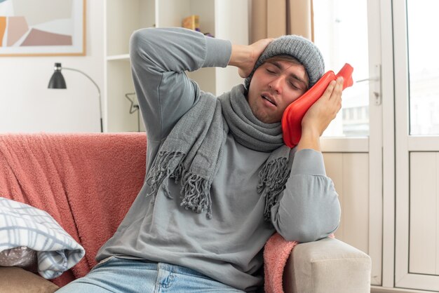 obolały młody chory mężczyzna z szalikiem na szyi w czapce zimowej kładący rękę na głowie i trzymający termofor siedzący na kanapie w salonie
