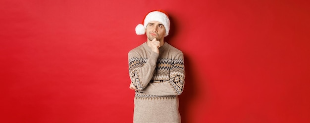 Bezpłatne zdjęcie obraz atrakcyjnego mężczyzny w świątecznym swetrze i santa hat myśli o prezentach noworocznych, patrząc na