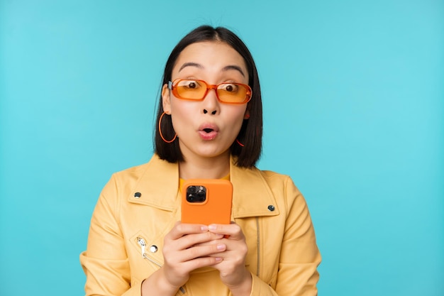 Obraz azjatyckiej dziewczyny w okularach przeciwsłonecznych wyglądającej na zdumioną i pod wrażeniem nagrywania wideo lub robienia zdjęć na smartfonie stojącej na niebieskim tle