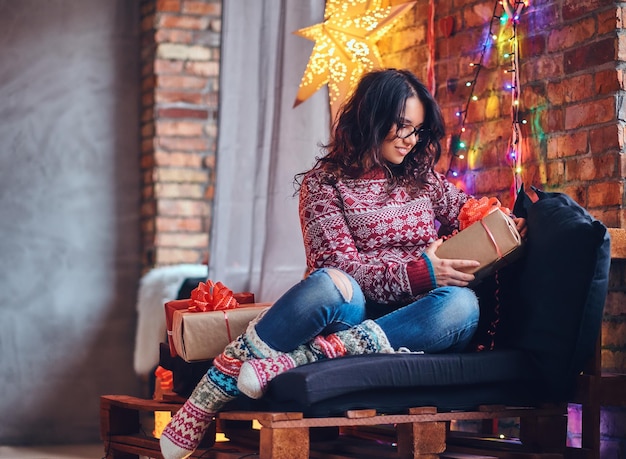 Obraz całego ciała brunetki kobiety w okularach, ubrany w dżinsy i czerwony sweter pozowanie na drewnianej kanapie w pokoju z dekoracją świąteczną.