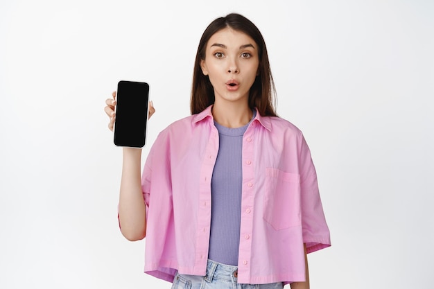 Obraz dziewczyny wygląda ciekawie, podczas gdy pokazuje ekran telefonu komórkowego przedstawia nową aplikację pokazującą białe tło aplikacji na smartfona
