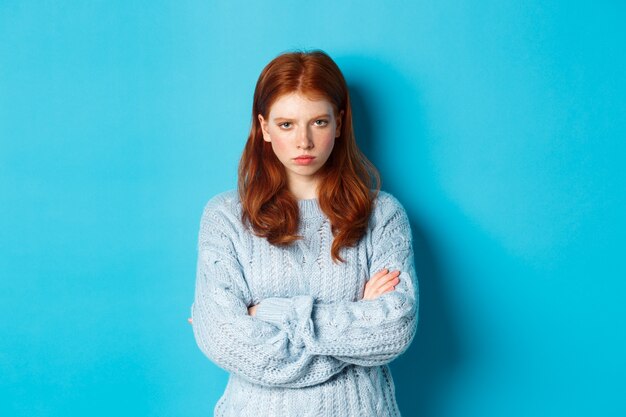 Obraz gniewnej rudej dziewczyny, która czuje się obrażona, skrzyżowane ręce na piersi i dąsa się, wpatrując się w szaloną kamerę, stojąc na niebieskim tle.