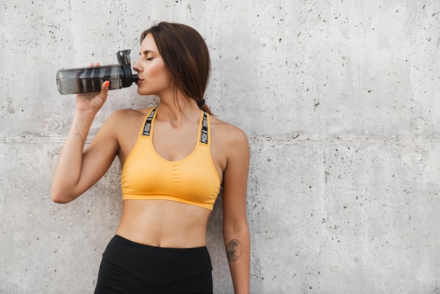 Obraz kobiety fitness w wodzie pitnej w odzieży sportowej z plastikowej butelki stojącej nad betonową ścianą na zewnątrz