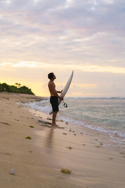 Obraz Krajobraz Mężczyzna Surfer Zajęty Chodzeniem Po Plaży O Wschodzie Słońca, Niosąc Deskę Surfingową Pod Pachą Z Falami Oceanu Załamującymi Się W Tle. Młody Przystojny Mężczyzna Surfer Na Oceanie