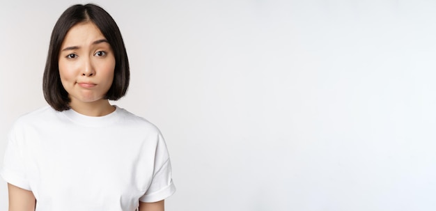 Bezpłatne zdjęcie obraz młodej azjatyckiej brunetki w białej koszulce uśmiecha się i patrzy z wątpliwościami, wahając się, stojąc na białym tle