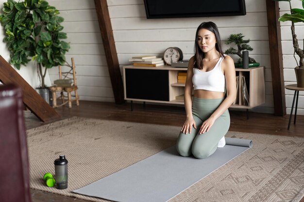 Obraz młodej azjatyckiej kobiety robiącej jogę w domu na macie podłogowej, medytującej w odzieży sportowej w salonie