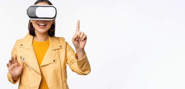 Obraz młodej azjatyckiej kobiety wearng VR headset używający okularów wirtualnej rzeczywistości i uśmiechający się, wskazując na coś, wybierając coś stojącego na białym tle