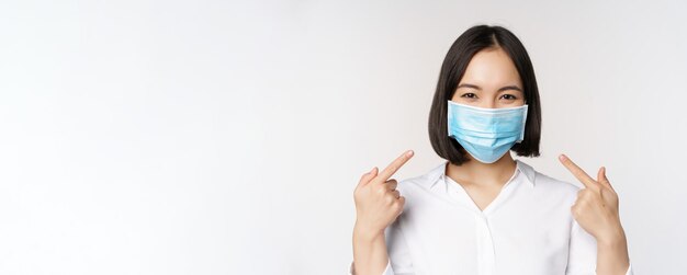 Obraz młodej azjatyckiej kobiety wskazującej na siebie podczas noszenia medycznej maski na twarz z koncepcją ochrony covid19 stojącej na białym tle