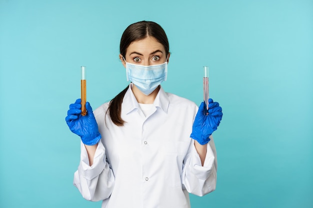 Obraz młodej lekarki, pracownika laboratorium prowadzącego badania, trzymającego probówki, noszącego maskę medyczną i gumowe rękawiczki, niebieskie tło