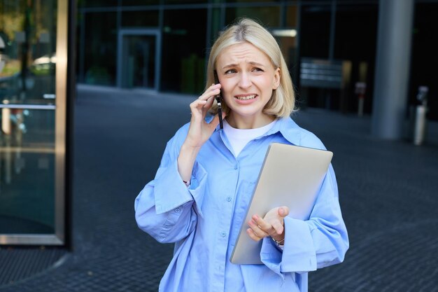 Bezpłatne zdjęcie obraz młodej uśmiechniętej kobiety pracującej w firmie stojącej w pobliżu biura z laptopem odpowiada na telefon