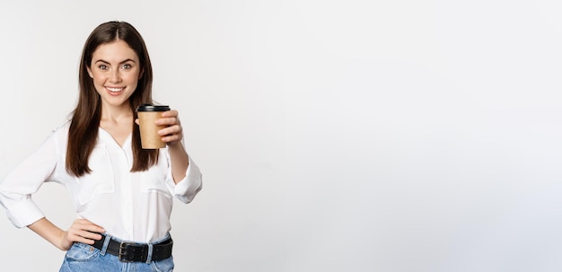 Obraz nowoczesnej kobiety biurowej trzymającej filiżankę kawy na wynos i uśmiechniętej stojącej na białym tle